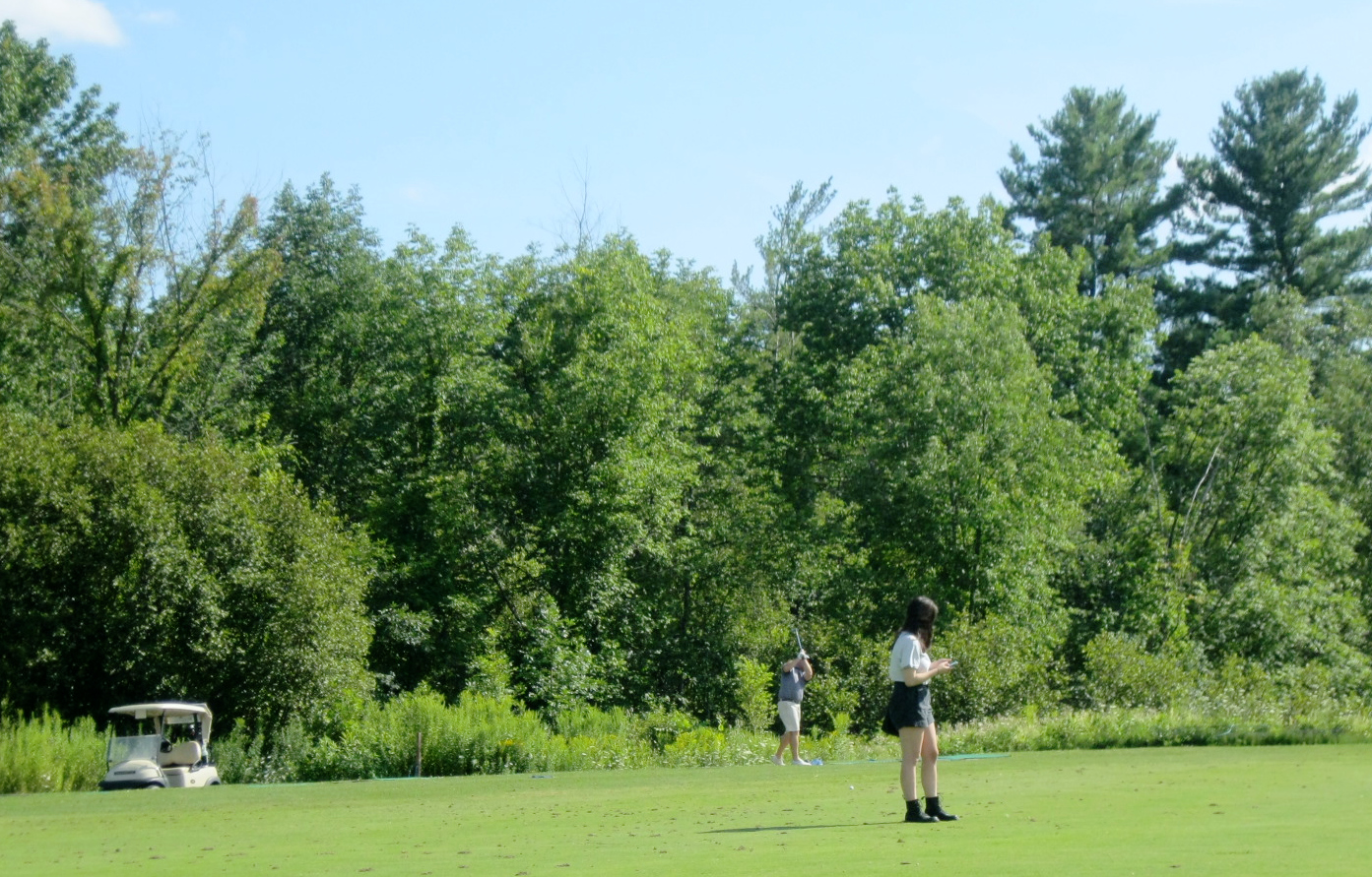 A young woman wearing black boots stands in the foreground while an older man in the background practices his golf swing.